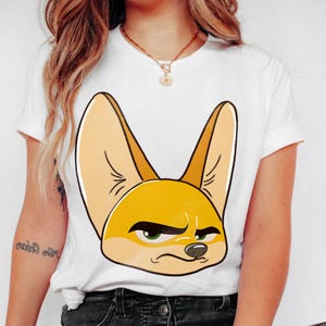 May include: White t-shirt featuring a cartoon illustration of a fennec fox with large ears and a slightly grumpy expression. The fox is primarily yellow and tan, with green eyes and a black outline. The shirt has short sleeves.