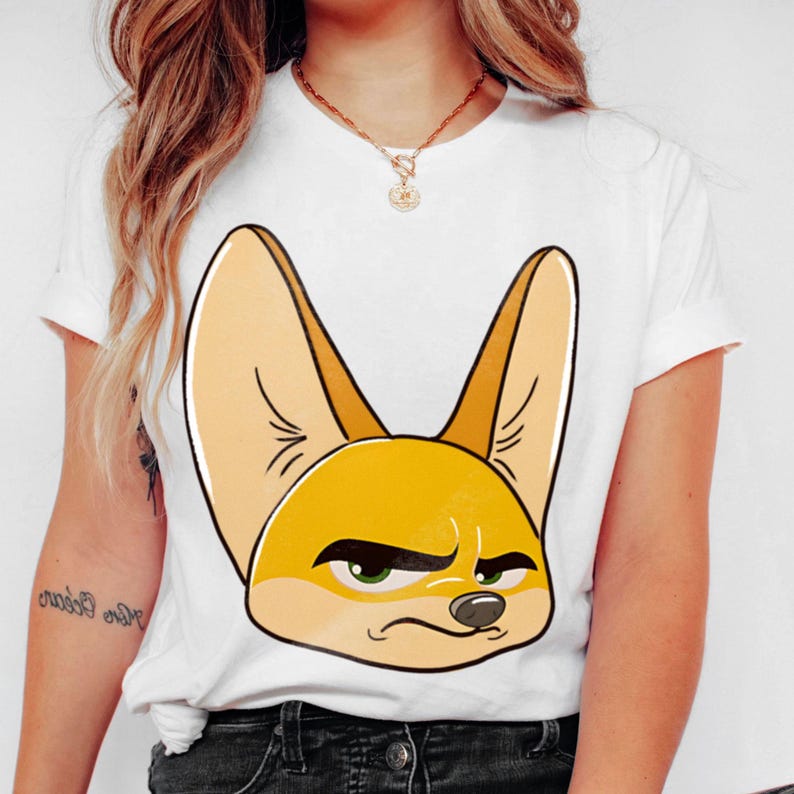 May include: White t-shirt featuring a cartoon illustration of a fennec fox with large ears and a slightly grumpy expression. The fox is primarily yellow and tan, with green eyes and a black outline. The shirt has short sleeves.