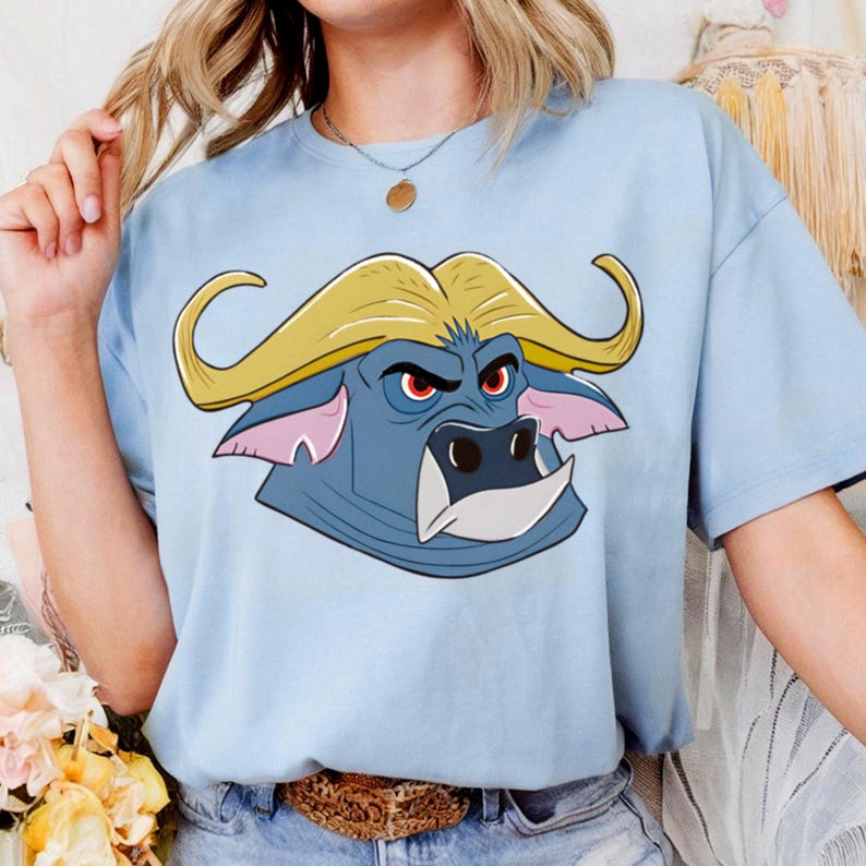 May include: Light blue t-shirt featuring a cartoon buffalo head with yellow horns, red eyes, and a white snout. The buffalo is set against a light blue background. The shirt is made of a soft material.