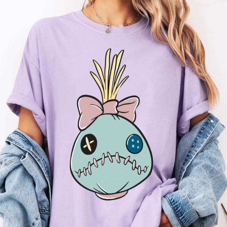May include: A light purple t-shirt featuring a cartoon-style doll head design. The doll head is light blue with a pink bow, yellow hair, and stitched details. The shirt has short sleeves and a relaxed fit.