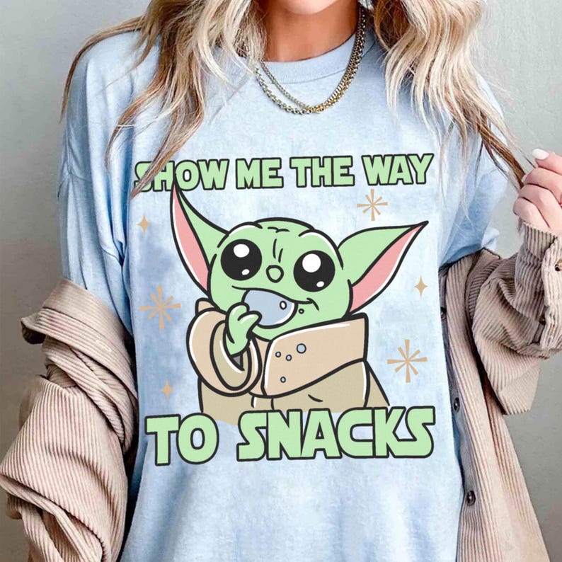 Peut inclure: T-shirt bleu clair avec un dessin anim&eacute; d'un personnage extraterrestre vert mangeant une collation. Le texte "SHOW ME THE WAY TO SNACKS" est imprim&eacute; en vert au-dessus et en dessous du personnage. Le t-shirt a des manches courtes.