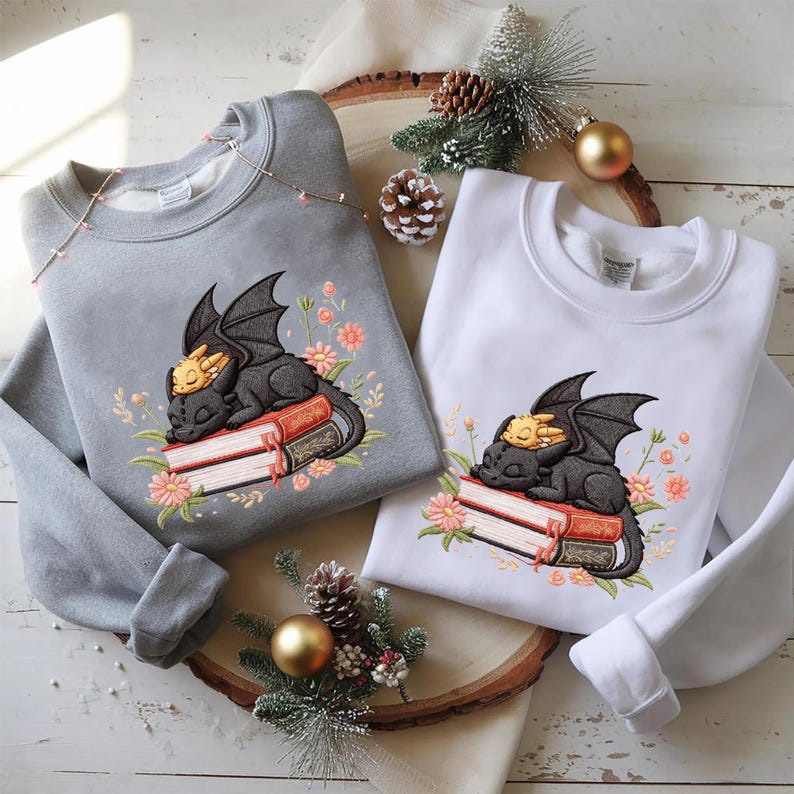 May include: Two sweatshirts, one gray and one white, each featuring an embroidered design of a black dragon sleeping on books, with a smaller yellow dragon on top. The design is surrounded by pink flowers.