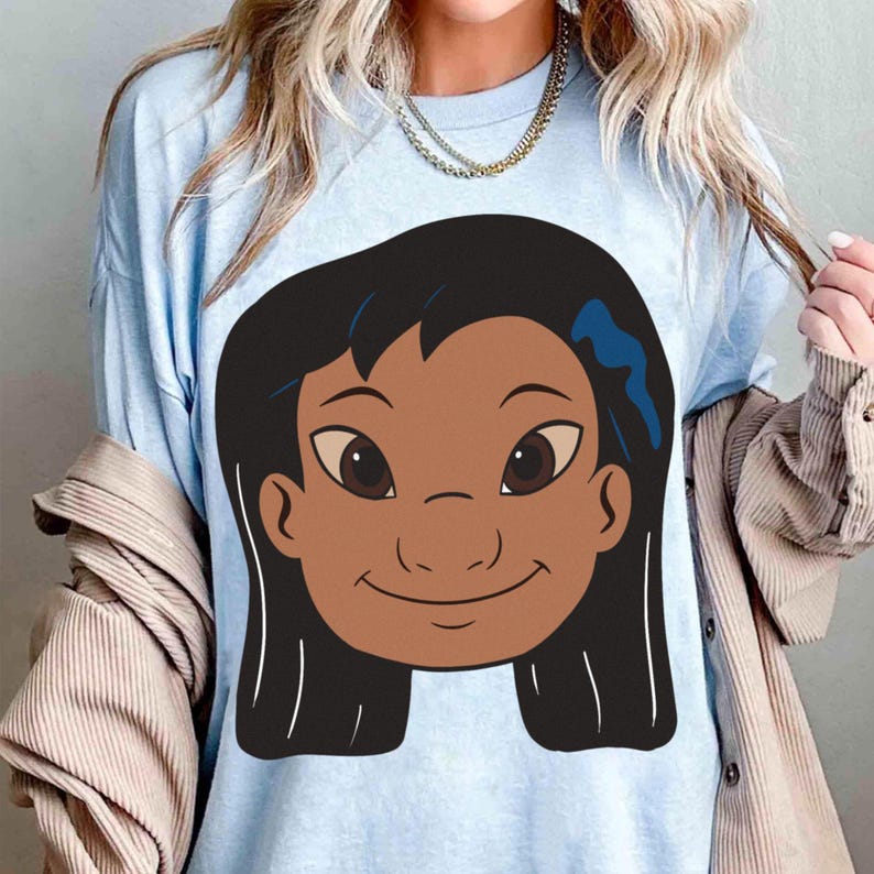 Puede incluir: Camiseta azul claro con una ilustraci&oacute;n de dibujos animados de una ni&ntilde;a sonriente con cabello oscuro y piel morena. El gr&aacute;fico est&aacute; centrado en la camiseta. La camiseta est&aacute; hecha de un material suave.