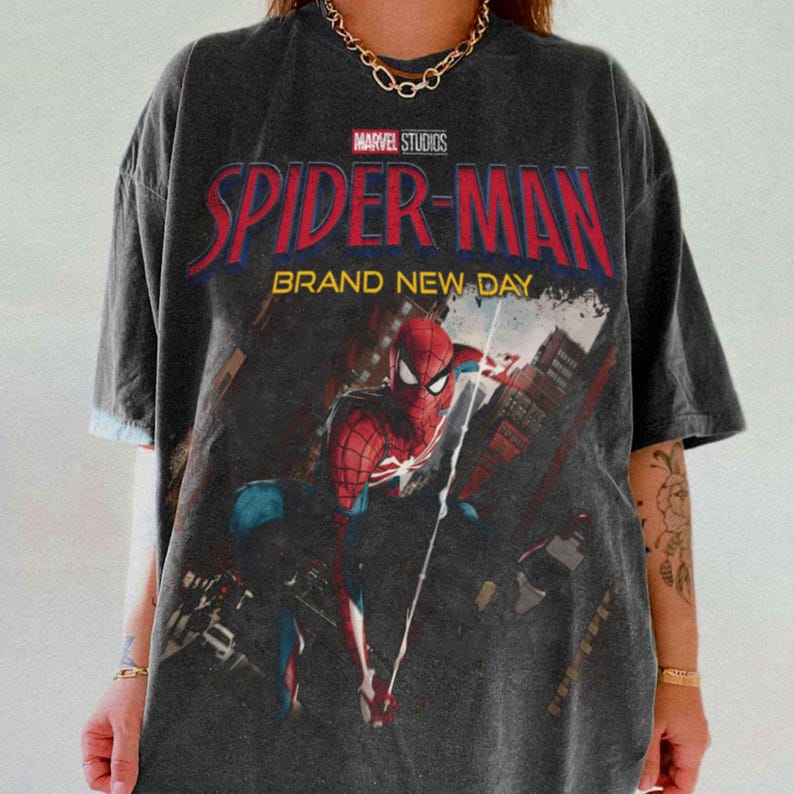 May include: Dark gray t-shirt featuring the Marvel Studios Spider-Man logo in red and blue, with the words "BRAND NEW DAY." The graphic shows Spider-Man in action against a cityscape backdrop. The shirt has a vintage, worn look.