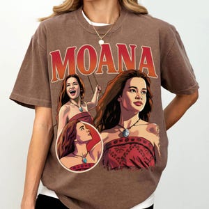 以下が含まれることがあります： モアナのキャラクターのグラフィックプリントが施された茶色の半袖Tシャツ。上部には大きな赤い文字でMOANAと書かれており、赤い柄のトップスとネックレスを身に着けたキャラクターの3つの異なるイラストが描かれています。