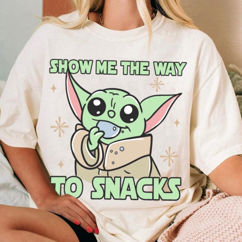 Peut inclure: T-shirt cr&egrave;me avec un personnage de dessin anim&eacute; vert tenant un objet bleu. Le texte "SHOW ME THE WAY TO SNACKS" est imprim&eacute; au-dessus et en dessous du personnage en lettres vertes. Des &eacute;toiles d&eacute;coratives sont &eacute;galement incluses.
