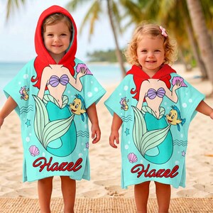 May include: Two children wearing light blue hooded beach towels with a mermaid design. The towels have red hoods, and the mermaid illustration features a purple seashell bra, red hair, and a teal tail. The name "Hazel" is written in red script.