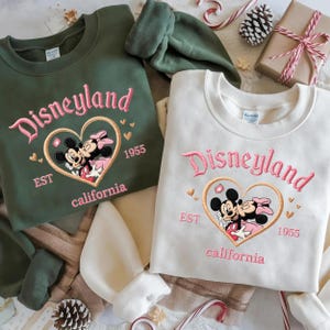 Peut inclure: Deux sweatshirts, un vert olive et un cr&egrave;me, brod&eacute;s avec "Disneyland" en rose. Chacun pr&eacute;sente un c&oelig;ur avec Mickey et Minnie Mouse, "EST 1955 California".