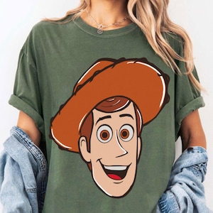 May include: Olive green t-shirt featuring a large cartoon graphic of a smiling cowboy character with a brown hat and brown hair. The shirt has short sleeves and a relaxed fit.