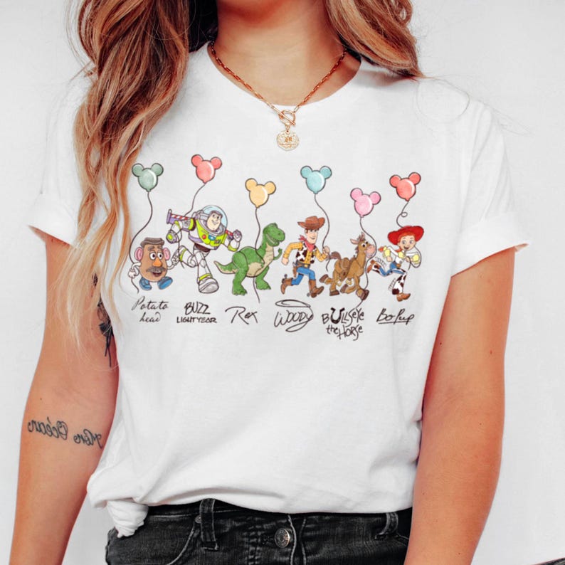 May include: White t-shirt with a vibrant illustration of Toy Story characters holding Mickey Mouse-shaped balloons. The design includes Potato Head, Buzz Lightyear, Rex, Woody, Bullseye the Horse, and Bo Peep. The characters' names are written below them.