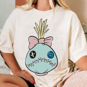 May include: Cream-colored t-shirt featuring a cartoon-style doll head design. The doll head is light blue with a pink bow, yellow hair, and stitched details. The design includes a black cross-shaped button and a blue button.