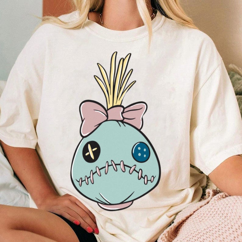May include: Cream-colored t-shirt featuring a cartoon-style doll head design. The doll head is light blue with a pink bow, yellow hair, and stitched details. The design includes a black cross-shaped button and a blue button.