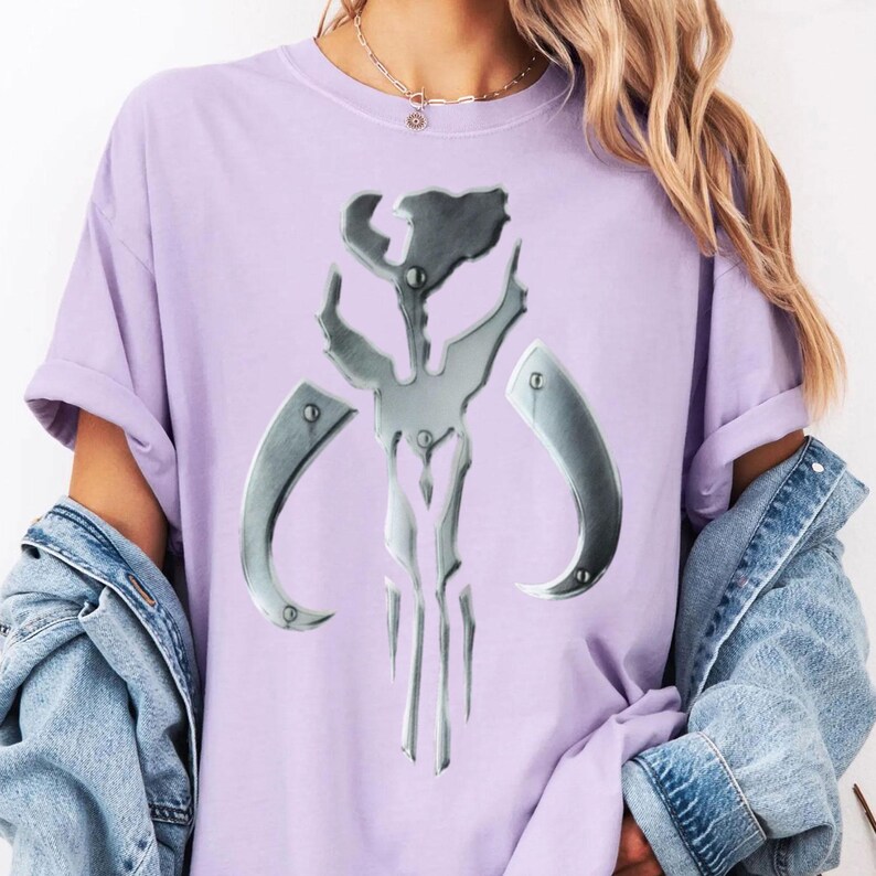 May include: A lilac t-shirt with a metallic silver emblem. The emblem is a symmetrical design with curved elements and rivets. The t-shirt has short sleeves and a relaxed fit.