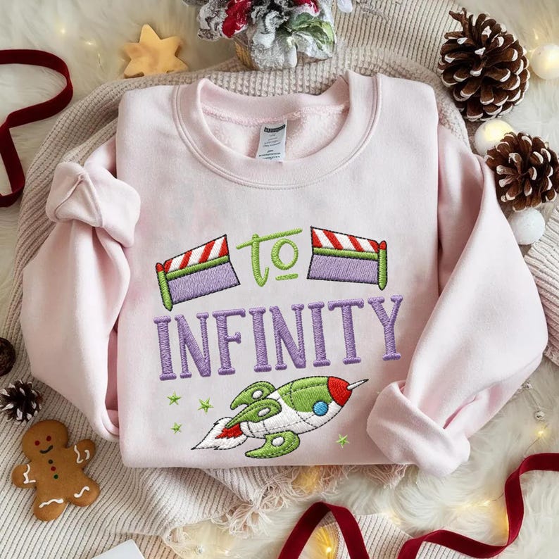 Puede incluir: Una sudadera rosa claro con la frase bordada "To Infinity" en morado y verde, junto con un dise&ntilde;o de cohete verde, rojo y blanco. La sudadera tiene mangas largas y cuello redondo.