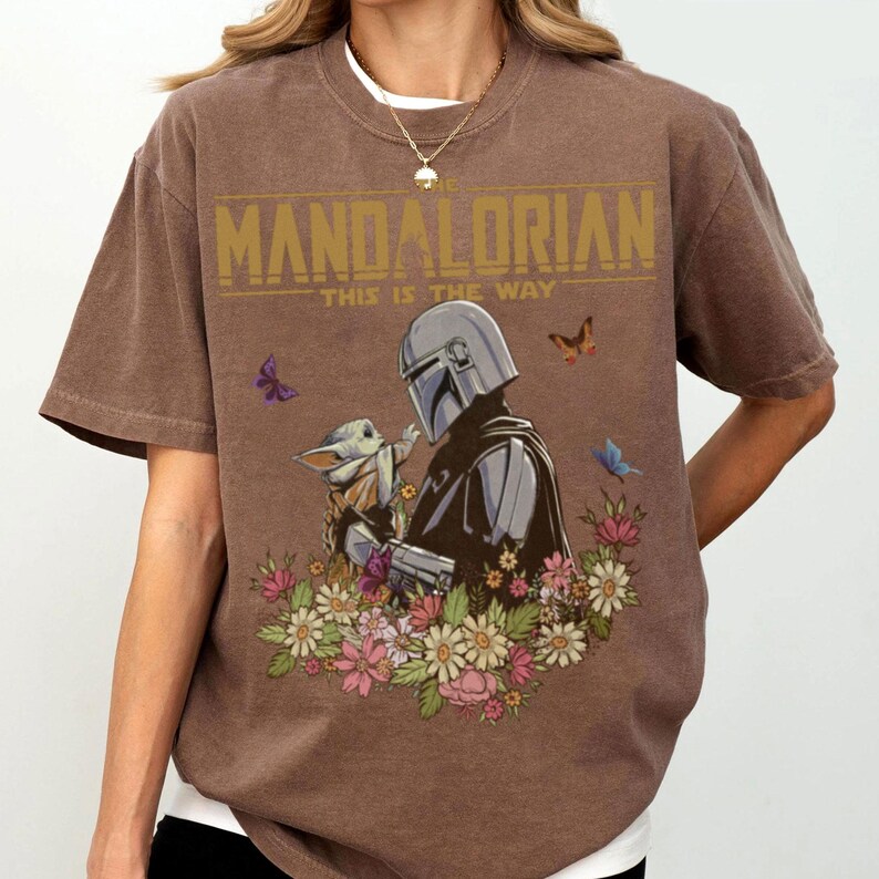 May include: Brown t-shirt featuring a graphic of The Mandalorian and Grogu surrounded by flowers and butterflies. The text "THE MANDALORIAN" and "THIS IS THE WAY" are printed in gold.