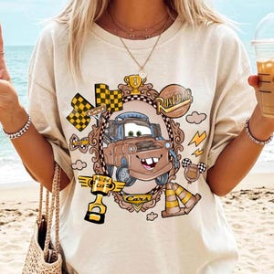 May include: Beige t-shirt featuring a cartoon image of Mater from the movie Cars, framed in a decorative oval. The design includes racing flags, a trophy, and the words "Rusteze" and "Cars".