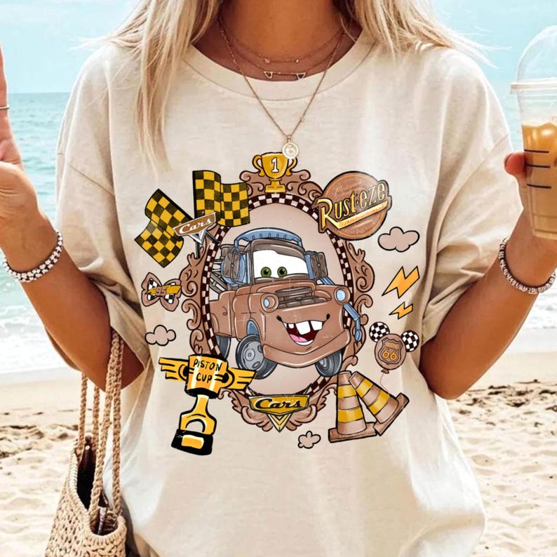 May include: Beige t-shirt featuring a cartoon image of Mater from the movie Cars, framed in a decorative oval. The design includes racing flags, a trophy, and the words "Rusteze" and "Cars".