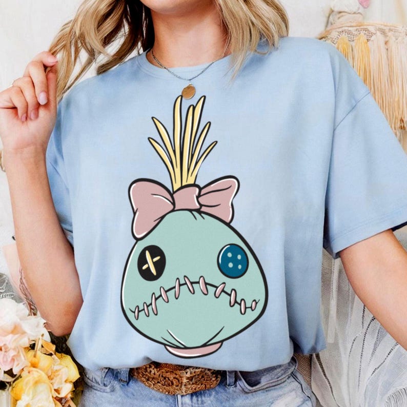 May include: Light blue t-shirt featuring a whimsical cartoon design. The graphic depicts a teal-colored sack-like character with a pink bow, stitched mouth, and button eyes. The character has yellow straw-like hair. The shirt is made of a soft material.