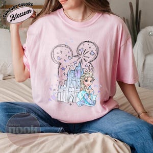 May include: A light pink t-shirt featuring a whimsical design. The graphic includes a castle, a princess, and a Mickey Mouse-shaped outline with stars and fireworks. The shirt has the text "Comfort Colors Blossom" at the top and "The Cozy Nook" at the bottom.