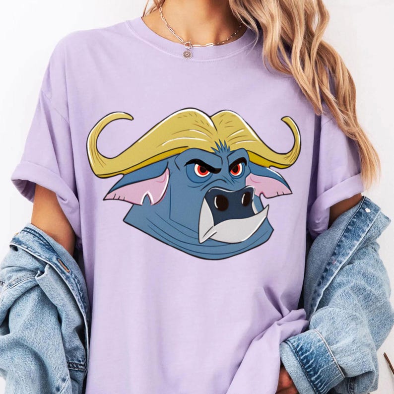 May include: A light purple t-shirt featuring a cartoon buffalo head with a blue face, yellow horns, and red eyes. The buffalo has a stern expression. The shirt has rolled-up sleeves and is paired with a denim jacket.