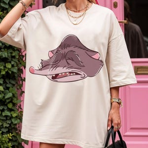 Peut inclure: T-shirt cr&egrave;me surdimensionn&eacute; avec un visage de possum de dessin anim&eacute; aux yeux ferm&eacute;s et &agrave; la bouche ouverte. Le possum est repr&eacute;sent&eacute; dans des tons de gris et de rose. Le t-shirt a des manches courtes.