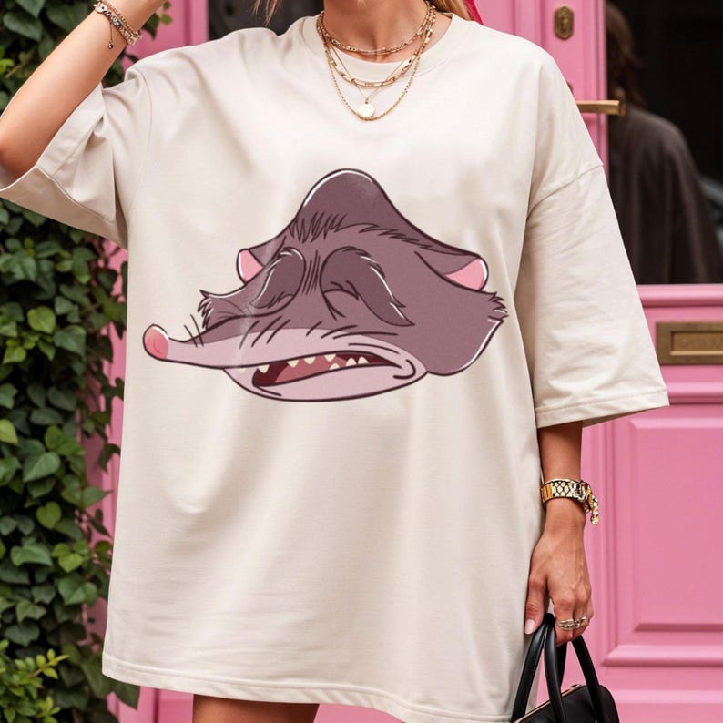 Peut inclure: T-shirt cr&egrave;me surdimensionn&eacute; avec un visage de possum de dessin anim&eacute; aux yeux ferm&eacute;s et &agrave; la bouche ouverte. Le possum est repr&eacute;sent&eacute; dans des tons de gris et de rose. Le t-shirt a des manches courtes.