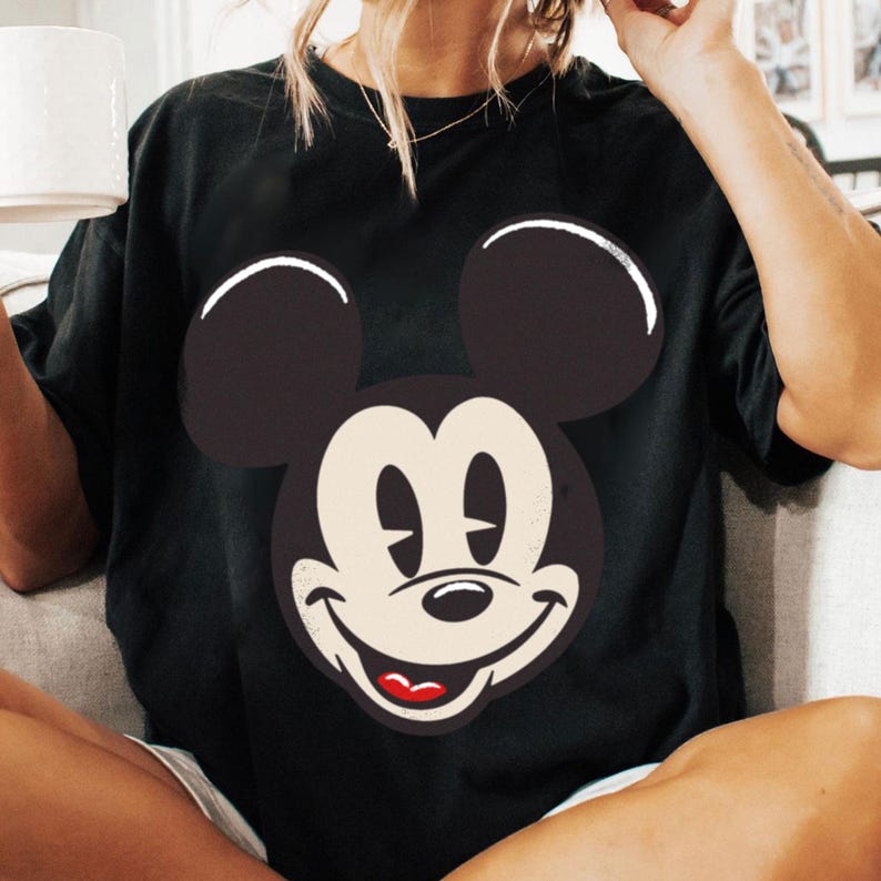 May include: Black t-shirt featuring a large graphic of Mickey Mouse's face. The design includes a smiling face with a cream-colored mouth and a red tongue. The ears are black with white outlines.