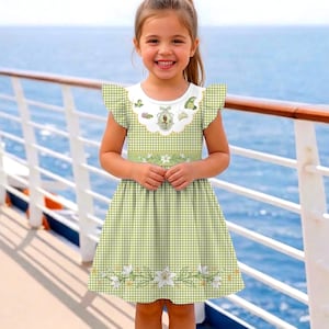 May include: A light green and white gingham dress with a white collar and flutter sleeves. The dress features a floral design on the hem and a decorative emblem on the collar. The dress is worn by a young girl.