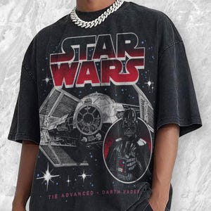 May include: Black t-shirt featuring a Star Wars graphic. The design includes the words "STAR WARS" in red and white, a Tie Advanced starfighter, and a portrait of Darth Vader. The text "TIE ADVANCED • DARTH VADER" is printed at the bottom.