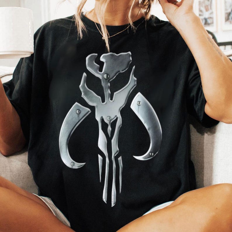 May include: Black t-shirt with a metallic silver Mandalorian skull emblem. The emblem is a stylized design with curved horns and a central vertical element. The shirt is short-sleeved and appears to be made of a soft material.
