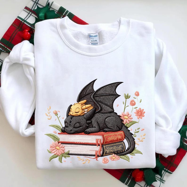 May include: White sweatshirt featuring an embroidered design of a black dragon and a yellow dragon resting on a stack of books. The design is surrounded by pink flowers and green leaves. The sweatshirt has long sleeves.