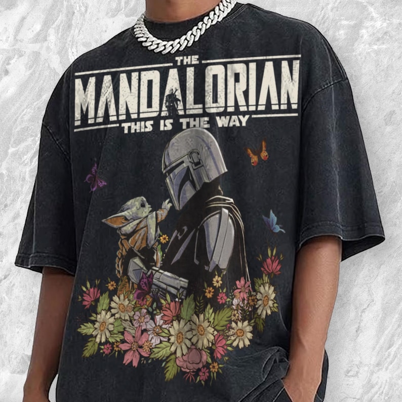 May include: Black t-shirt featuring a graphic of The Mandalorian and Grogu surrounded by flowers and butterflies. The text "THE MANDALORIAN THIS IS THE WAY" is printed across the top. The shirt has a vintage, distressed look.