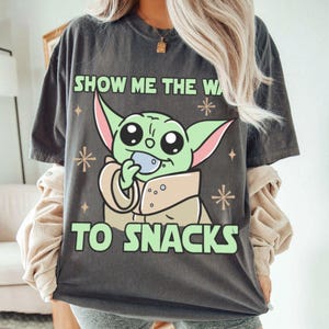 Peut inclure: T-shirt gris fonc&eacute; avec un personnage de dessin anim&eacute; vert tenant un petit objet bleu. Le texte "SHOW ME THE WA" et "TO SNACKS" est imprim&eacute; en vert. Le design comprend des &eacute;toiles et des diamants. V&ecirc;tement d&eacute;contract&eacute;.