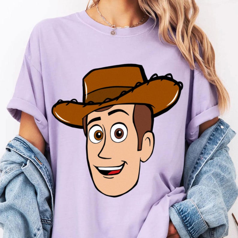 May include: A light purple t-shirt featuring a cartoon illustration of Woody from Toy Story. The character has brown hair, a brown cowboy hat, and a smiling expression. The shirt has short sleeves.