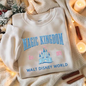 May include: Cream-colored sweatshirt with the words "MAGIC KINGDOM" embroidered in blue, and a castle graphic. Below the castle is the text "WALT DISNEY WORLD". The sweatshirt also has the text "Est. 1971".