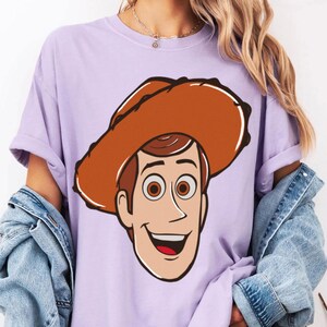 May include: A light purple t-shirt featuring a cartoon illustration of Woody from Toy Story. The character has brown hair, brown eyes, and a cowboy hat. The design is centered on the shirt.