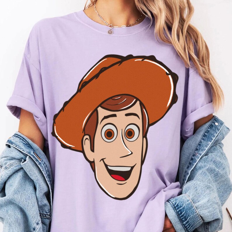 May include: A light purple t-shirt featuring a cartoon illustration of Woody from Toy Story. The character has brown hair, brown eyes, and a cowboy hat. The design is centered on the shirt.