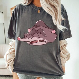 Peut inclure: T-shirt gris fonc&eacute; avec un dessin de visage de possum de dessin anim&eacute; en nuances de violet et de rose. Le possum a les yeux ferm&eacute;s, un long museau et des dents visibles. Le t-shirt a des manches courtes et une coupe d&eacute;contract&eacute;e.