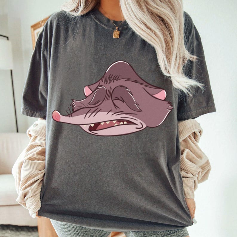 Peut inclure: T-shirt gris fonc&eacute; avec un dessin de visage de possum de dessin anim&eacute; en nuances de violet et de rose. Le possum a les yeux ferm&eacute;s, un long museau et des dents visibles. Le t-shirt a des manches courtes et une coupe d&eacute;contract&eacute;e.