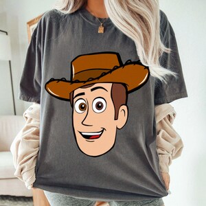 May include: Dark gray t-shirt featuring a cartoon illustration of a cowboy character with a brown hat, brown hair, and a smiling face. The design is centered on the front of the shirt. The shirt has short sleeves and a relaxed fit.