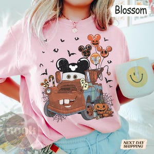 May include: Pink t-shirt with a Halloween-themed graphic featuring a cartoon truck with Mickey Mouse ears, balloons, pumpkins, and bats. The word "Blossom" is printed on the top right.