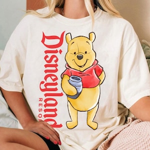 May include: Cream-colored t-shirt featuring a cartoon illustration of Winnie the Pooh in a red hoodie, holding a blue honey pot. The words "Disneyland Resort" are printed vertically in red on the left side of the shirt.