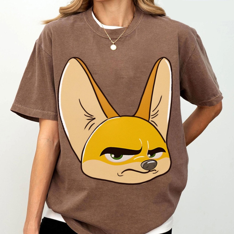 May include: Brown t-shirt featuring a cartoon fennec fox with a grumpy expression. The fox has large ears, a yellow face, and green eyes. The shirt has short sleeves and a crew neck.