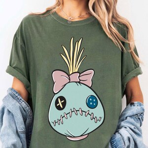 May include: Olive green t-shirt featuring a cartoon design of a teal doll-like figure with a pink bow, yellow hair, and stitched details. The design includes a black cross and a blue button for eyes, and a stitched mouth.
