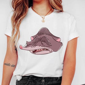 Peut inclure: T-shirt blanc avec un dessin de possum de dessin anim&eacute;. Le possum est repr&eacute;sent&eacute; dans des tons gris et roses, avec les yeux ferm&eacute;s et la bouche ouverte. Le t-shirt a des manches courtes et un col rond.