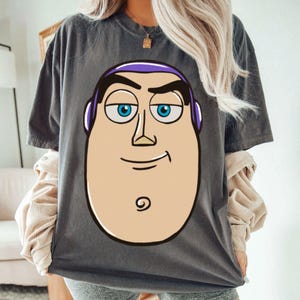 May include: Dark gray t-shirt featuring a large cartoon image of Buzz Lightyear's face. The graphic includes his blue eyes, purple headpiece, and a smiling expression. The shirt is a casual, oversized style.