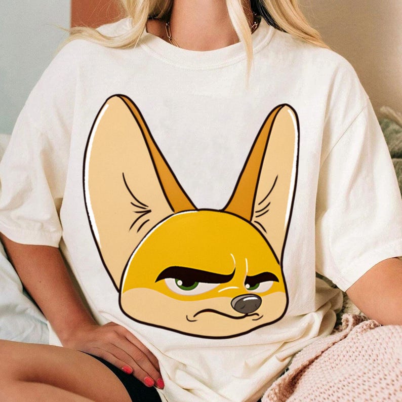 May include: Cream-colored t-shirt featuring a large cartoon illustration of a fennec fox with an annoyed expression. The fox has large ears, yellow fur, and green eyes. The shirt is a casual, unisex style.