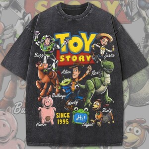 Pode incluir: Uma t-shirt de manga curta cinzento-carv&atilde;o com uma impress&atilde;o gr&aacute;fica das personagens de Toy Story, incluindo Buzz, Jessie, Bullseye, Alien, Woody, Rex, Hamm e Slinky. O texto diz Toy Story, Since 1995 e Hi num quadrado verde.