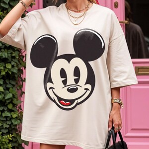 May include: Cream-colored oversized t-shirt featuring a large graphic of Mickey Mouse's face. The design includes black ears, a smiling face with white eyes, and a red mouth. The shirt has short sleeves.