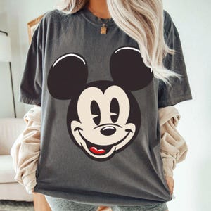 May include: Dark gray t-shirt featuring a large graphic of Mickey Mouse's face. The design includes black ears, a white face, and a smiling expression with a red mouth. The shirt has short sleeves and a relaxed fit.